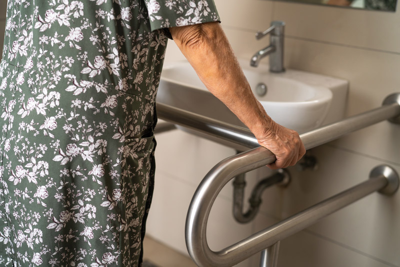 Cómo ayudar a una persona mayor a ir al baño de forma segura Mujer mayor apoyándose en un asidero de seguridad junto al lavabo